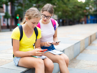 Two female students use phones to check lessons and write down completed assignments in a notebook