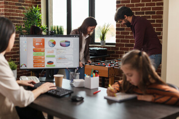 Agency business people playing table soccer while young woman working sitting at computer. Workplace coworkers enjoying leisure time together while having a table soccer competition in workspace.