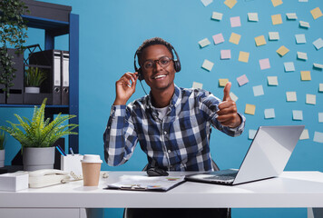 Cheerful smiling african american helpline center operator wearing headset, showing thumbs up approval sign while on call with client. Joyful customer support representative sitting at desk in office.