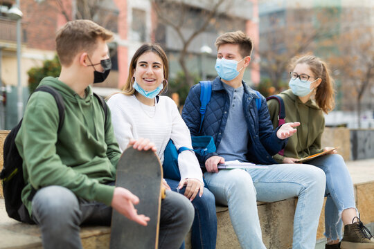 Smiling teenager boys and girls in protective face masks talking with each other sitting on stairs on city street
