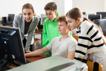 Obraz premium Teenage boy sitting at table and using computer during computer science lesson. His teacher and classmates standing and looking at monitor.