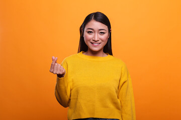 Cheerful friendly smiling asian woman demanding money and rewards by showing hand cash gesture on orange background. Happy woman smiling on payday while rubbing fingers and showing money gesture.