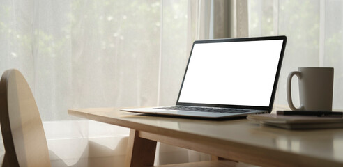 Laptop computer with blank screen, coffee cup and book on wooden table in bright living room.