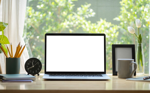 Laptop computer with blank display, coffee cup, pencil holder and picture frame on wooden table in bright living room.