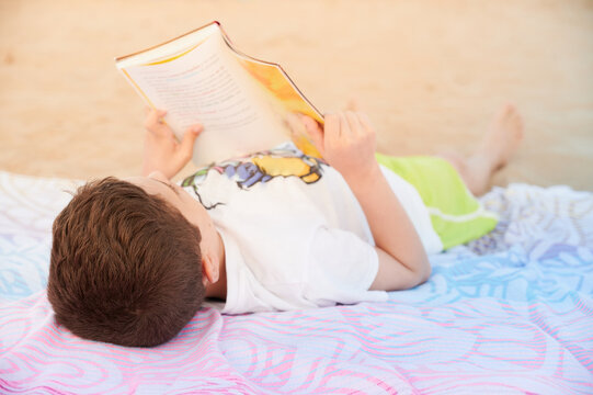 Elementary school age boy lying on a towel at the beach, reads totally focused an adventure book on summer vacation. Summertime lifestyle concept.