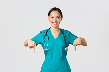 Covid-19, healthcare workers, pandemic concept. Smiling pleasant asian female doctor, therapist or physician in scrubs with stethoscope, pointing fingers down, show clinic banner