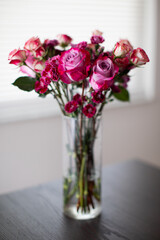 bouquet of roses, tulips, carnations in a clear vase on a wooden table in front of a window at the home