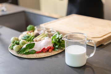 Coconut cream ,milk, is contained in a large measuring cup beside a group of food ingredients and these products is in front of a wood cutting board.
