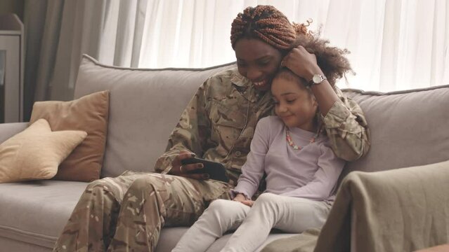 Slowmo of happy young Black woman in military uniform embracing her little daughter sitting together on couch in living room, watching video on smartphone and smiling, reuniting after long separation