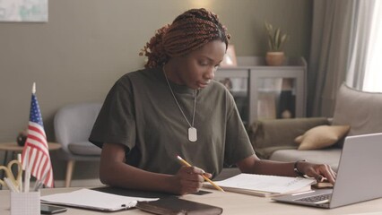 Waist up slowmo of young Black military woman with dreadlocks in khaki t shirt using laptop and examining documents while working from home at desk with us flag on it