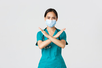 Covid-19, coronavirus disease, healthcare workers concept. Serious displeased, frowning asian doctor in scrubs show cross gesture in disapproval, stop someone, prohibit action, white background