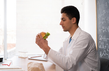 Young teacher having breakfast with hamburger at workplace, lunch in classroom with junk cheeseburger, student in white coat indoors eating fast food, nutrition
