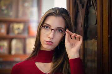 Young girl in the glasses stands next to a bookcase.