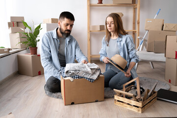 a young couple in love in a new apartment on the day of the move sit on the floor unpack things. The couple moves into a new apartment. Lots of packing boxes around them.
