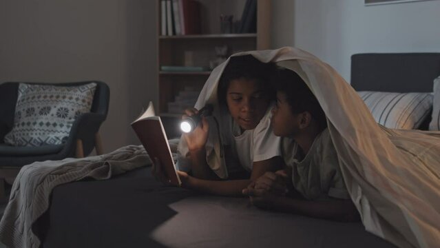 Medium of happy African American little siblings lying on stomachs under blanket on bed at night, reading book with flashlight, smiling and talking