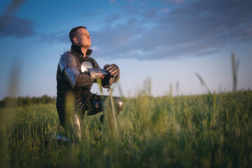 Medieval knight kneels in the field with his hand on the sword on a dark sky background.