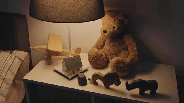 High-angle view of stuffed bear, various wooden toys and lit lamp on bedside table at night