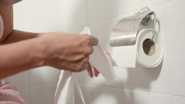 Closeup hand pulling toilet paper roll in holder for wipe, woman sitting on toilet she taking and tearing white tissue on wall to towel clean in bathroom, Healthcare concept