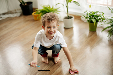 The boy is squatting on the floor in the room, holding a marker in his hand, wiping the floor with a napkin