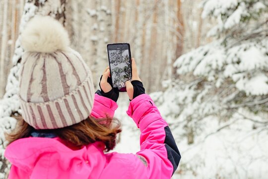 The Girl Takes Pictures Of Nature On A Smartphone. A Teenager In Bright Warm Overalls Takes A Photo Of A Winter Forest On A Cell Phone Camera