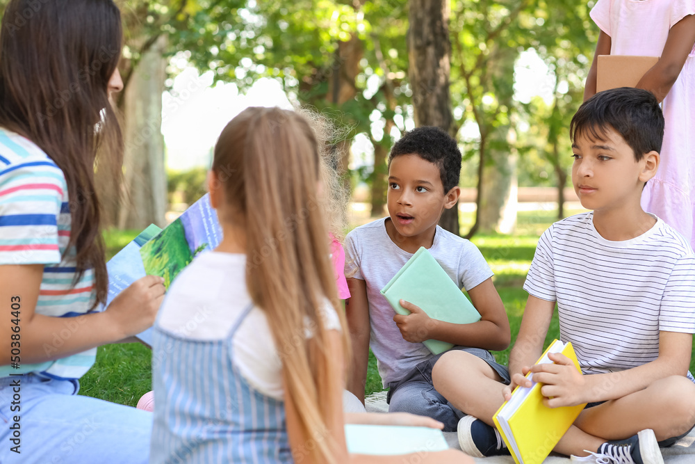 Wall mural Teacher reading book to her little pupils in park