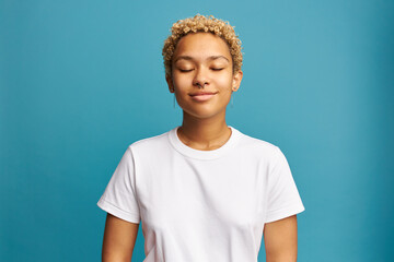 Head and shoulders image of charming adorable female of 20s in white mockup t-shirt with copy space, standing over blue background with closed eyes, enjoying smelling fresh air, doing deep breath