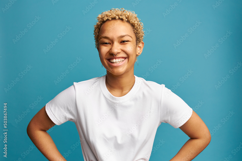 Wall mural Horizontal image of cheerful overjoyed female with lovely blonde curls in white mockup t-shirt isolated over blue background with copy space for your promotional content. Human emotions and feelings