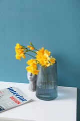 Vase with yellow flowers, decor and magazine on table near blue wall