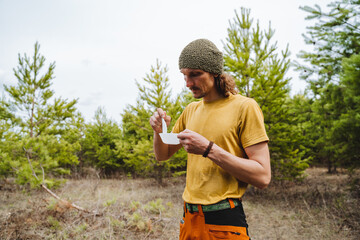 A guy eats food from a plate in nature, camping in the forest, a tourist man dine on porridge on a hike, a man in a yellow T-shirt stands against the background of the forest, a hipster with a beard