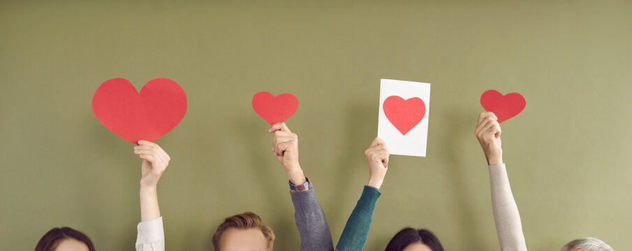 Unidentified men and women with raised hands hold models of hearts. Paper red hearts of different sizes in hands of unrecognizable people on khaki background. Concept of love, charity and volunteering