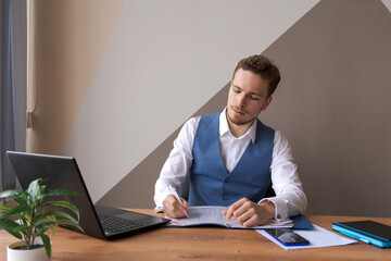 Business man works in an office with laptop and documents on his desk in shirt and waistcoat with a...