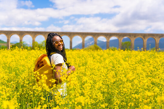 Portrait Of A Black Ethnic Girl With Braids Looking At The Sun, Sunglasses, Traveler, In A Field Of Yellow Flowers