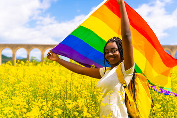 A smiling black ethnic girl with braids holding the LGBT flag in a field of yellow flowers, pride day, lesbian free women concept - Powered by Adobe