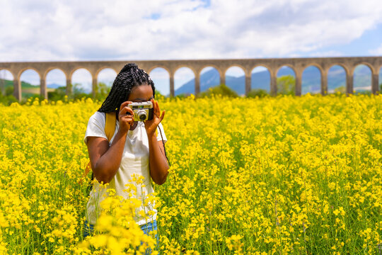 Taking photos with a vintage camera, a black ethnic girl with braids, a traveler, in a field of yellow flowers