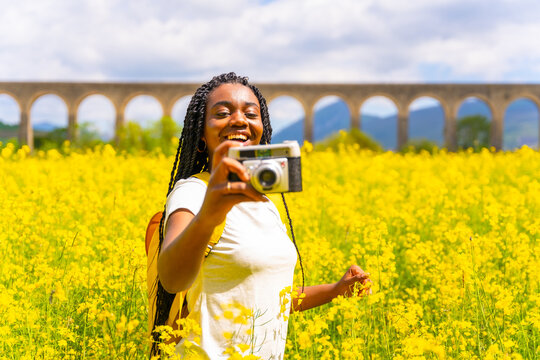 Taking photos smiling with a vintage camera, a black ethnic girl with braids, a traveler, in a field of yellow flowers
