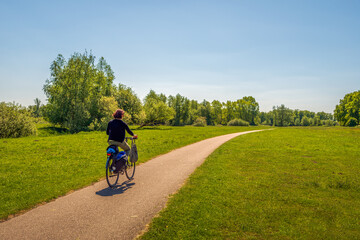 Woman cycles alone on a narrow cycle path in a rural area with grass, trees and bushes. It is a warm and sunny day in spring. The photo was taken near the Dutch city of Breda, North Brabant.