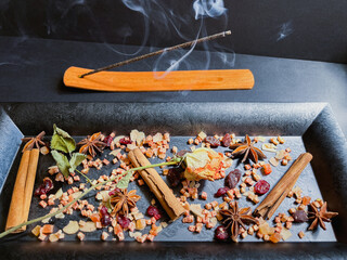 A mixture of dried fruits and spices lies on a black tray next to a burning incense stick. Anise stars with cinnamon and raisin sticks. food background.