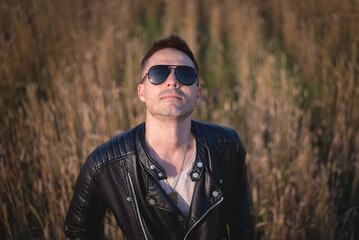 A man in the leather black jacket and sunglasses looks up the sky among the wheat field.
