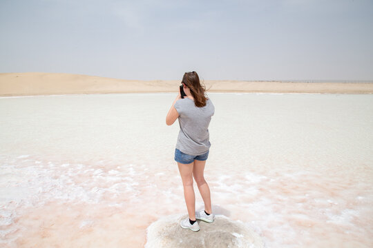  girl tourist standing on a rock taking photos in the desert with salt pan with pink water with grain and out of focus