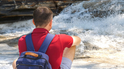 Back view of a male traveler in a red polo sitting by a roaring mountain river. Man alone travels...