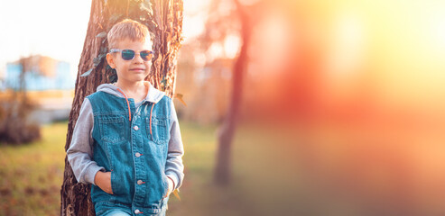 A lonely boy with glasses is standing near a tree. There is a park in the background. Copy space. Web banner. The concept of child disability, autism and psychology