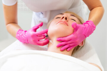 Anti-aging beauty injections. An adult blonde woman at a cosmetologist's appointment. A doctor in pink medical gloves injects the medicine into the client's neck