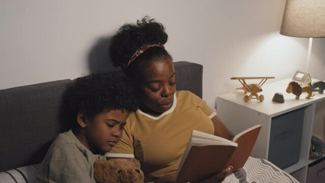 Medium of young African American mother reading book to cute nine-year-old son, lying in bed at night, boy holding toy bear and sleeping