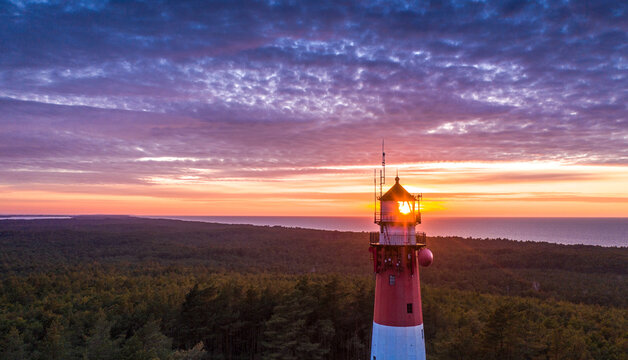 Beautiful sunset over green forest. View of the lighthouse, sea in the background. Orange sun, pastel pink clouds.
