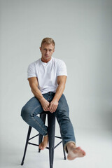 muscular male model actor in a studio in jeans and a white T-shirt posing sitting on a chair. Acting tests casting.