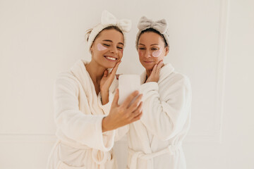 Happy caucasian young ladies in white coats and headbands take pictures on phone. Blondes get rid of dark circles around eyes with patches. Beauty treatments, morning ritual
