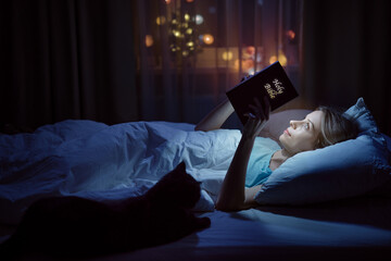 Magic book. Young woman reading Bible at cozy bed with her cat.