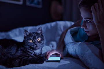 Young woman with her cat using smartphone in bed in her cozy bedroom at night.