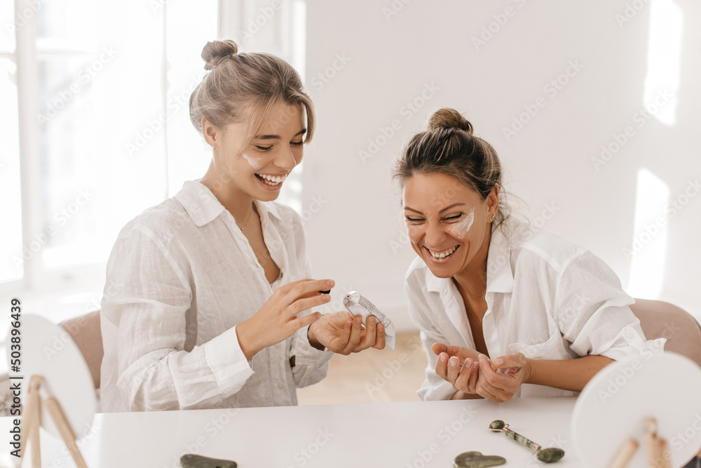 Wall mural Happy young caucasian ladies in good mood use face cream sitting in studio. Blondes wear white shirts laugh together. Day with skin care and health.