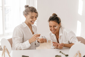 Obraz premium Happy young caucasian ladies in good mood use face cream sitting in studio. Blondes wear white shirts laugh together. Day with skin care and health.